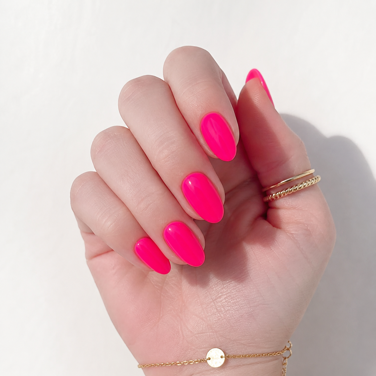 Close-up of fair, cool-toned hand with vivid neon pink gel nails, almond shape, gold rings, and bracelet on a clean white background