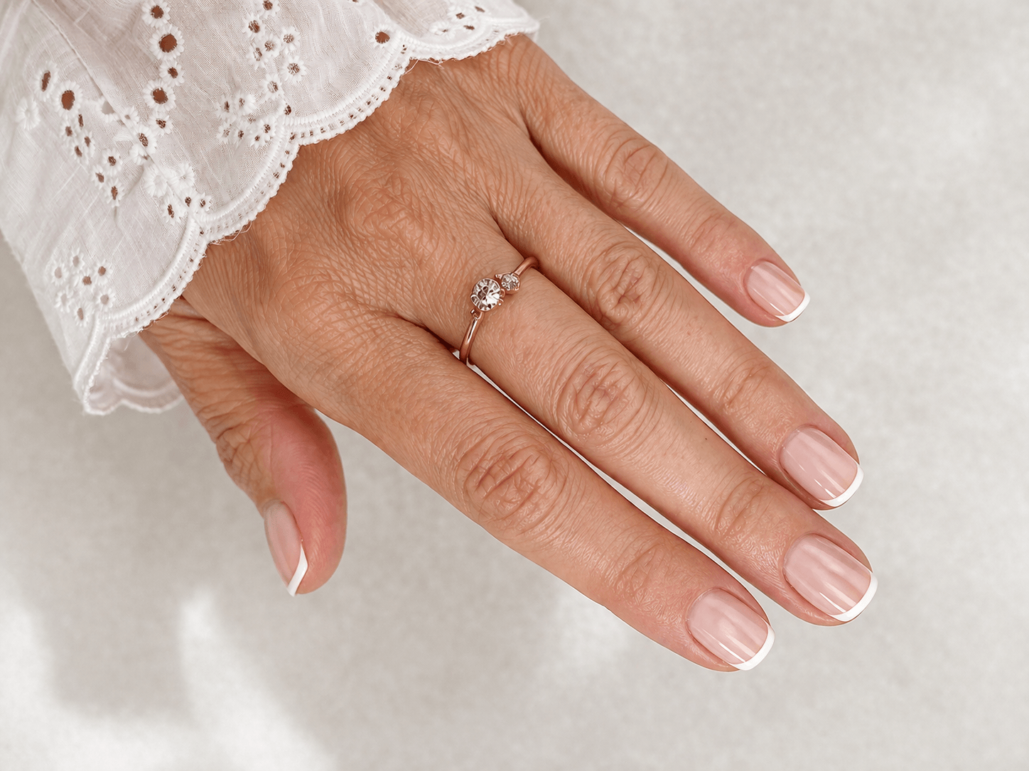 Close-up of mature hand with short squoval nails in sheer nude gel and subtle micro French tips, photographed indoors with a softly blurred neutral background
