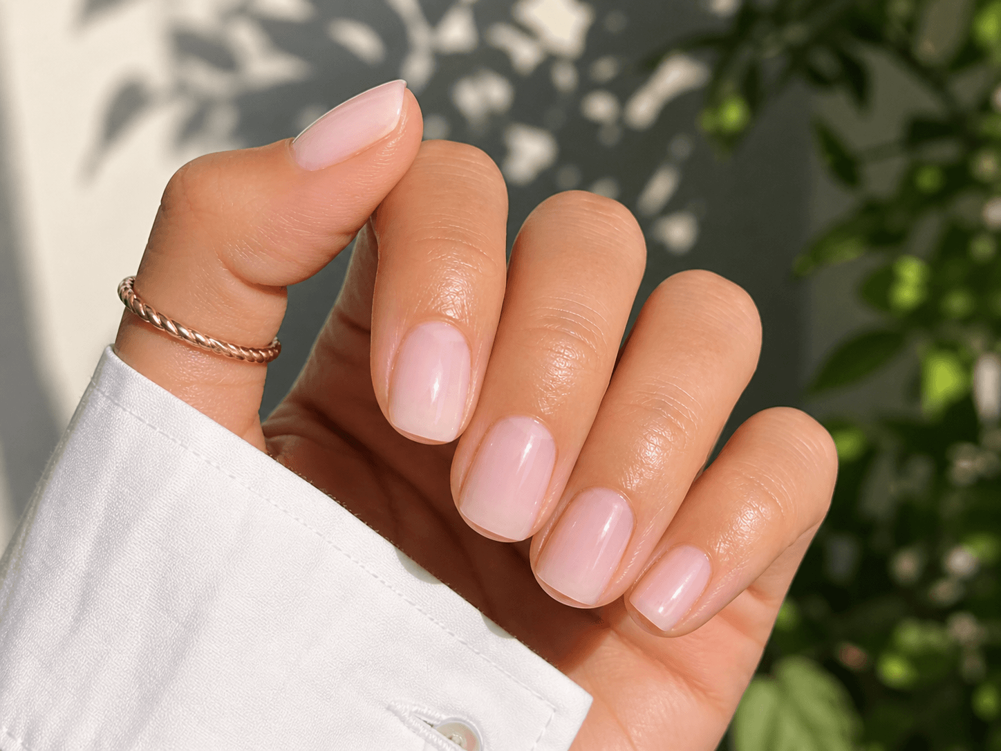 Close-up of hand with short squoval nails in sheer milky pink gel, wearing a white shirt sleeve and gold thumb ring in natural daylight