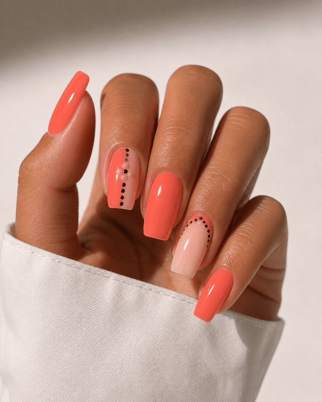 Close-up of tan hand with warm coral gel nails featuring minimal dot nail art in a glossy finish on a clean background