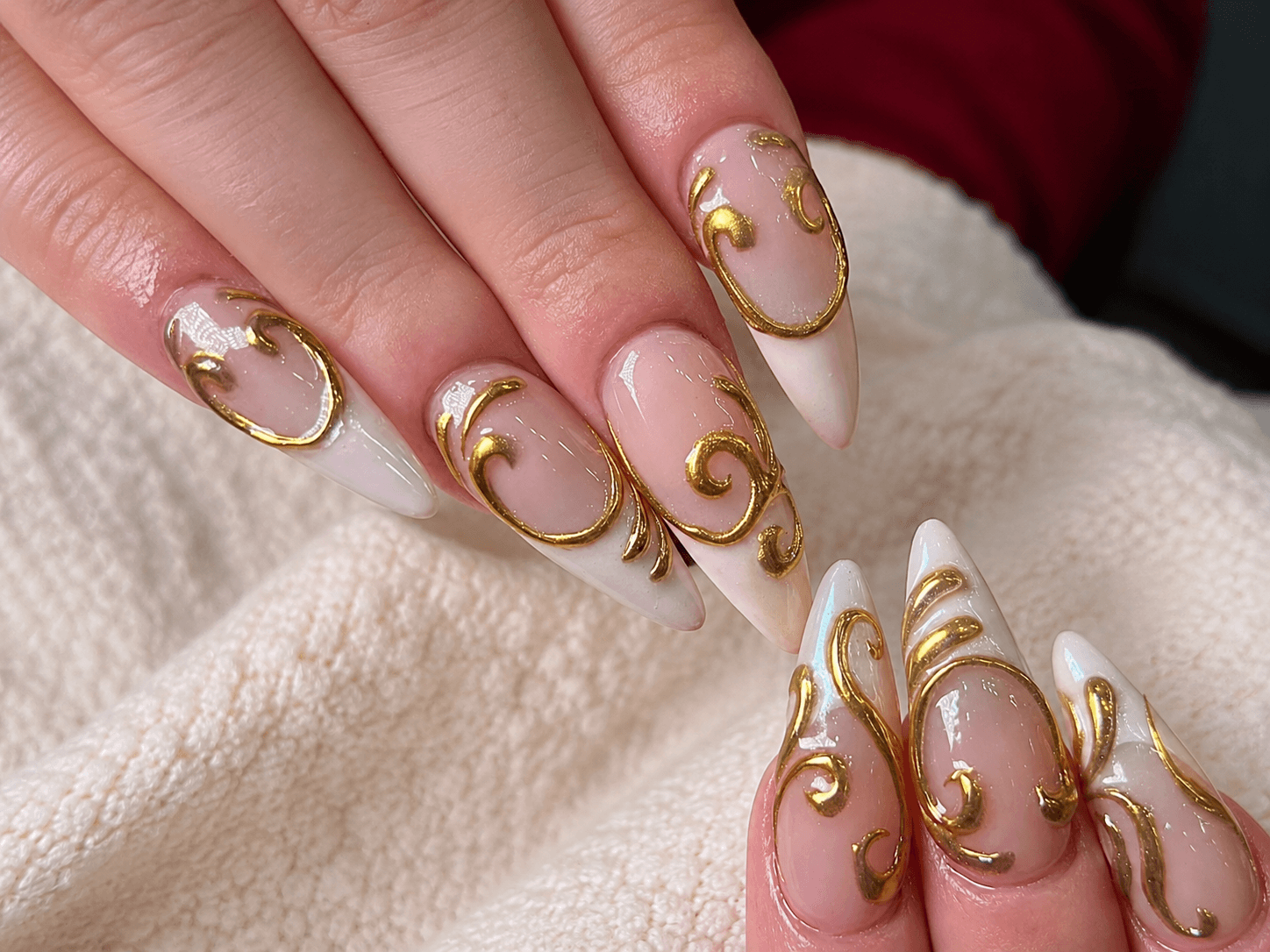 Elegant nude and white stiletto nails with gold swirl nail art on a soft cream fabric background