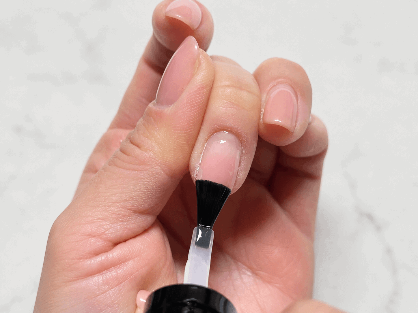Close-up of applying regular base nail polish over natural gel nails with a brush on a marble background